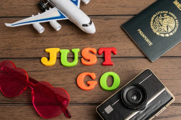 Airplane, passport, pink heart glasses, vintage camera and the phrase just go made from colorful letters on wooden background