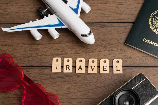 Airplane, pink heart glasses, vintage camera, passport and the word travel made from wooden letters on a wooden background