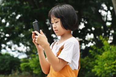 Parkta cep telefonu kullanan mutlu Asyalı kız çocuğu. Çocuk gülümsüyor ve ekrandaki cihazlara bakıyor. İletişim kavramı, teknoloji.