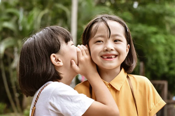 Two little sister girls whisper in ear at park outdoor.