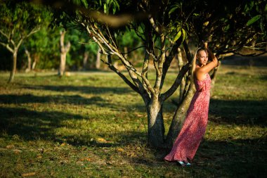 Tropik ağaç yakınındaki pembe elbiseli günbatımında poz güzel genç kız
