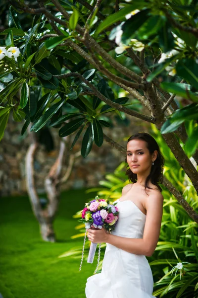 Beautiful young bride posing in wedding dress with tropical tree holding wedding bouquet