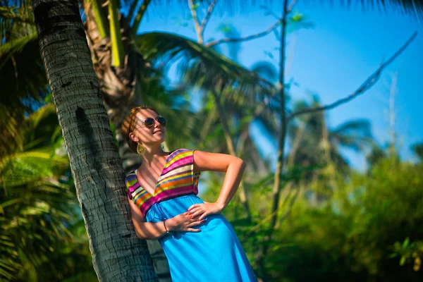 Güzel moda genç kadın elbise ve güneş gözlüğü palmiye ağaçları ve gökyüzü bir arka plan üzerinde bir ağaca yaslanmış