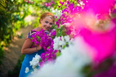 Çiçekler ortasında duran genç güzel kadın. Kız mavi bir elbise giymiş, mor ve beyaz çiçekler tarafından çevrilidir.