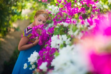 Genç güzel kadın güzel çiçek kokulu. Kız mavi bir elbise giymiş, mor ve beyaz çiçekler tarafından çevrilidir.