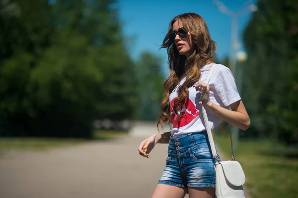 Een aantrekkelijk meisje in zonnebrillen en denim shorts wacht in het Park — Stockfoto