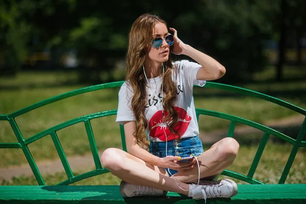 Genç güzel kız parkta bir bankta oturan kulaklıklar müzik dinlemek.