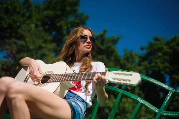 美丽的年轻时髦女孩弹吉他户外在公园里坐在长凳上 — 图库照片