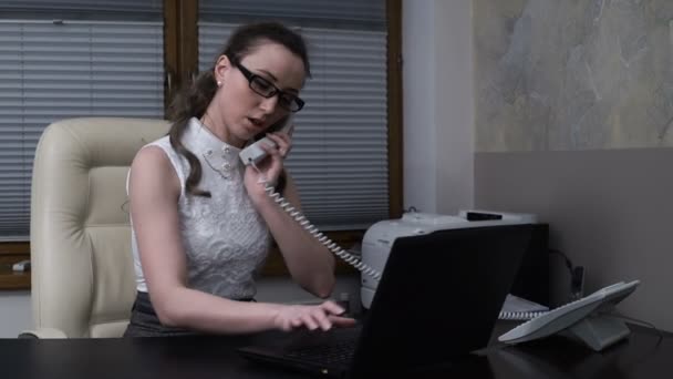 Mujer joven trabajando en el ordenador portátil y hablando por teléfono — Vídeos de Stock