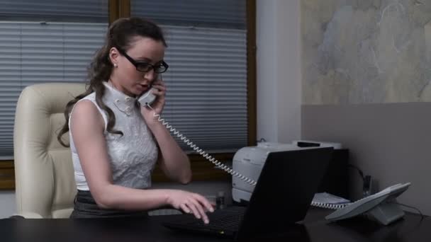 Молода жінка працює на ноутбуці і розмовляє по телефону — стокове відео