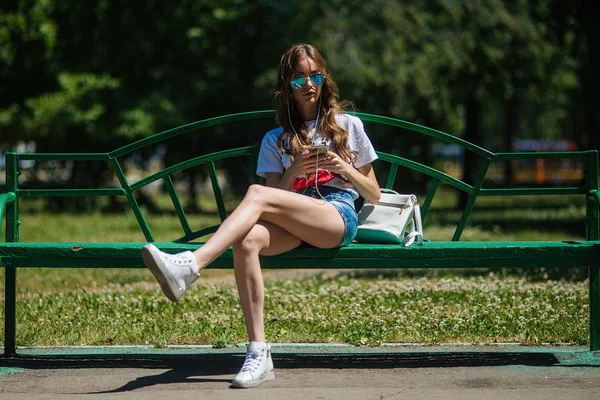 Genç güzel kız parkta bir bankta oturan kulaklıklar müzik dinlemek.