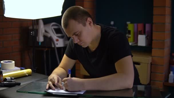 El joven dibuja un boceto sobre la mesa para tatuajes. El tipo dibuja un cuadro en papel — Vídeo de stock