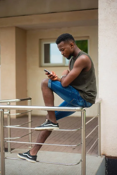 Hipster, un joven que usa un smartphone. Adolescente afroamericano sosteniendo un teléfono inteligente móvil sentado en la barandilla — Foto de Stock