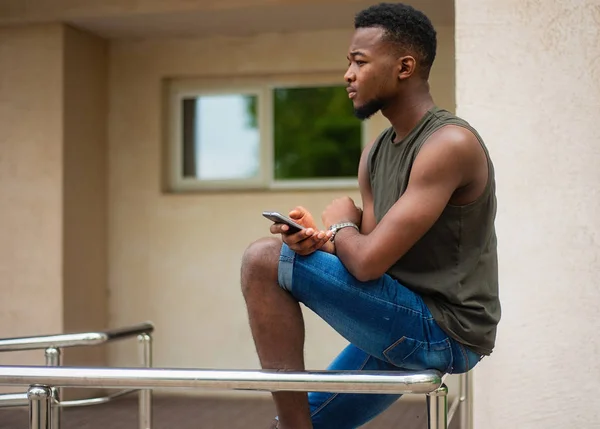 Hipster, un joven que usa un smartphone. Adolescente afroamericano sosteniendo un teléfono inteligente móvil sentado en la barandilla — Foto de Stock