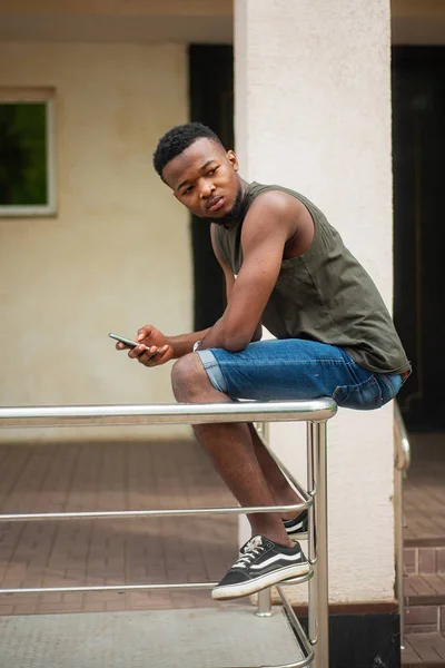 Hipster, un joven que usa un smartphone. Adolescente afroamericano sosteniendo un teléfono inteligente móvil sentado en la barandilla — Foto de Stock