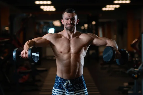 Handsome young muscular man trains his shoulders with dumbbells in the gym