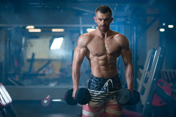 Güzel genç kaslı adam dumbbells ile el spor salonunda poz
