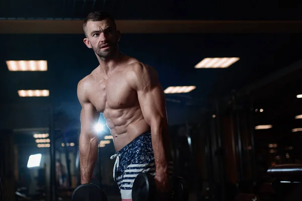 Beautiful young muscular man posing with dumbbells in hand at the gym