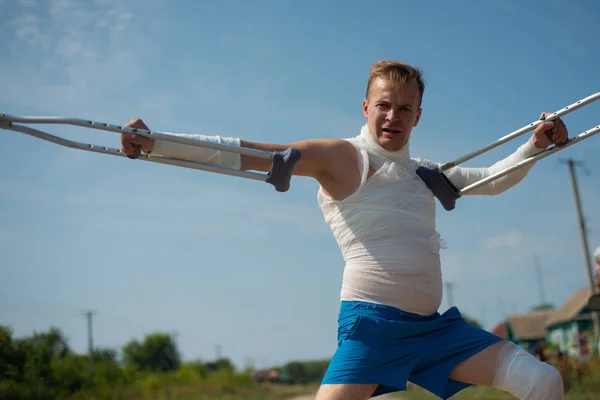 A young man in bandages on crutches walking on the road in the countryside, having fun and making grimaces