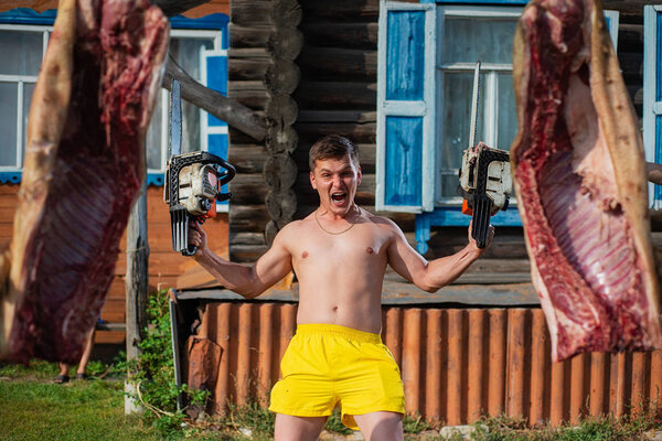 the man screams terribly, holding two chainsaws in his hands against the background of pig carcasses