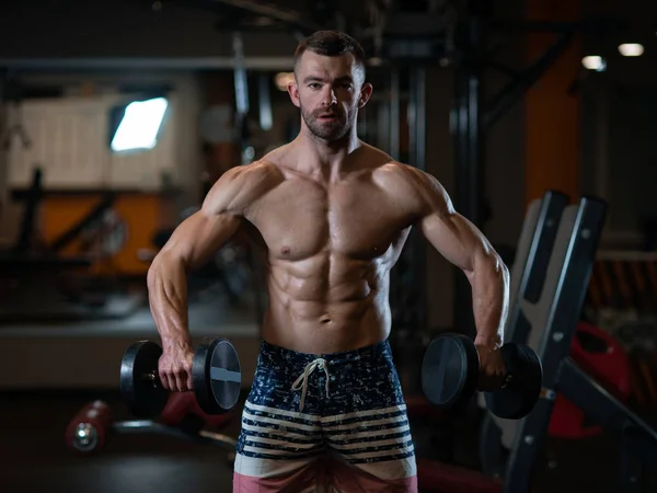 Handsome young muscular man trains his shoulders with dumbbells in the gym