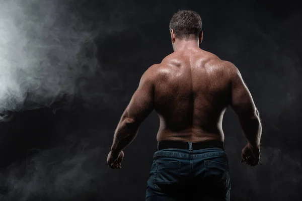 Powerful muscular man posing on black background showing his back in smoke