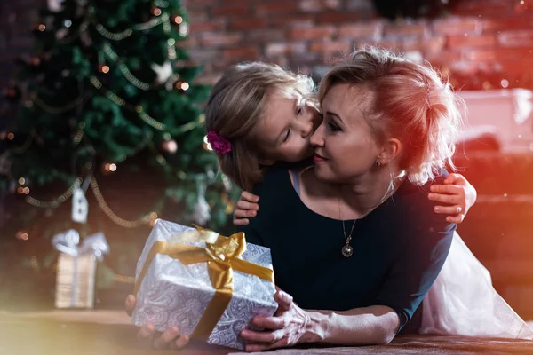 Mom gave her daughter a gift for the new year, and the daughter kisses her mother on the background of the Christmas tree