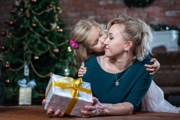 Mom gave her daughter a gift for the new year, and the daughter kisses her mother on the background of the Christmas tree