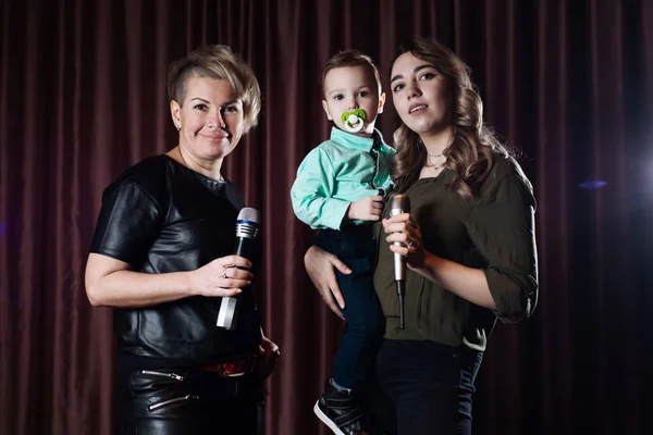 Two women and a small child sing on stage in microphones in karaoke on a background of red curtains