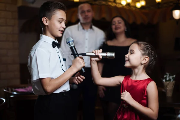 Little brother and sister sing karaoke songs in microphones, and their parents sing in the back