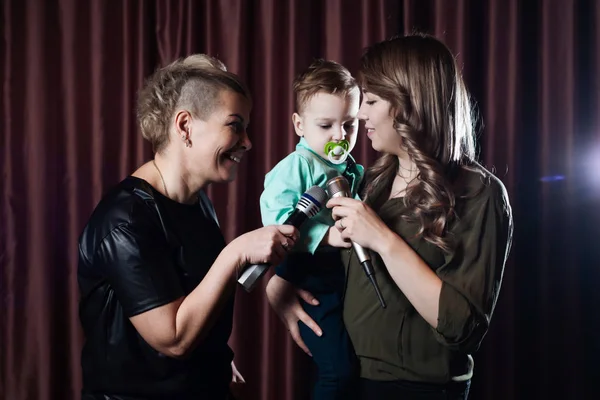 Two women and a small child sing on stage in microphones in karaoke on a background of red curtains