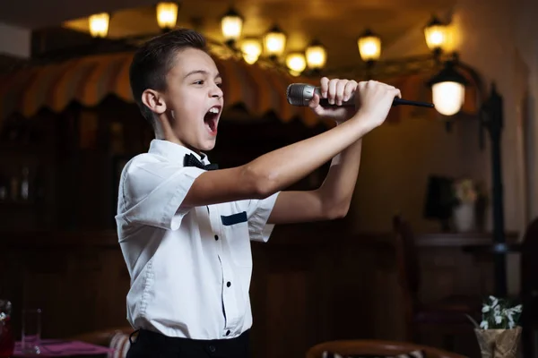 Pojke i en vit skjorta sjunger i mikrofonen i en restaurang. Baby karaoke. — Stockfoto