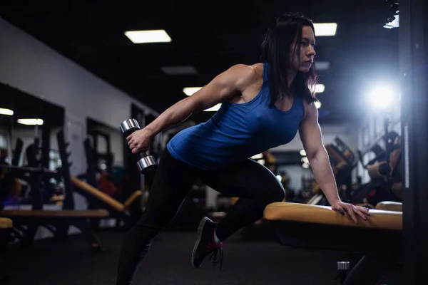 Sports fitness girl trains back muscles and biceps with dumbbells in the gym