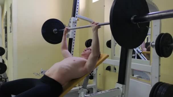 Joven musculoso en press de banca. Guy entrena en el gimnasio con una barra — Vídeos de Stock