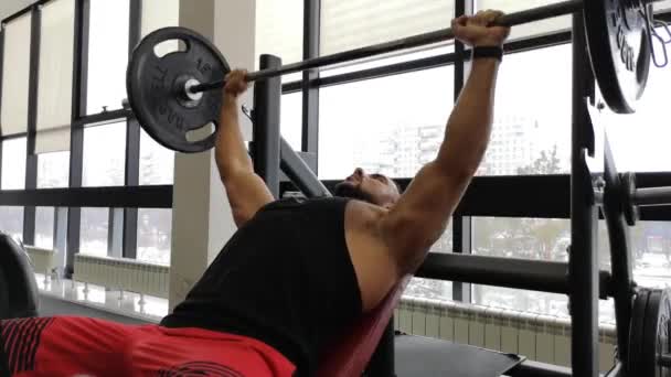 Joven musculoso en press de banca. Guy entrena en el gimnasio con una barra — Vídeo de stock