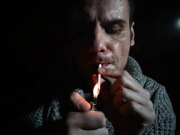 Foto escura e enrugada de um jovem fumando em um fundo preto. Um homem acende um cigarro — Fotografia de Stock