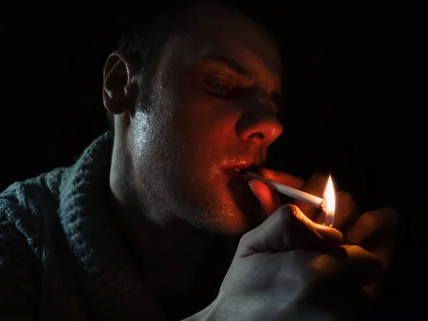 Foto escura e enrugada de um jovem fumando em um fundo preto. Um homem acende um cigarro Imagens De Bancos De Imagens Sem Royalties