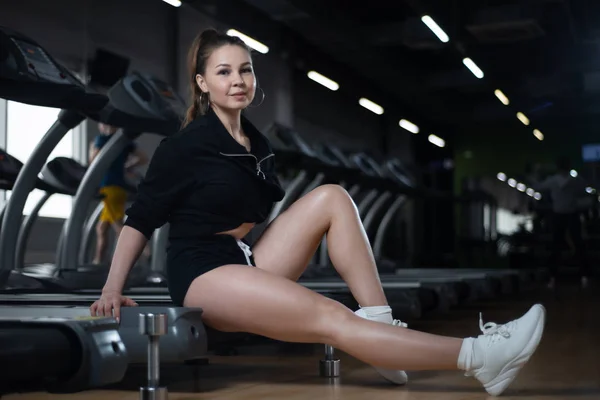 Фитнес-девушка позирует в спортзале сидя на беговой дорожке, хвастаясь своими яйцами — стоковое фото