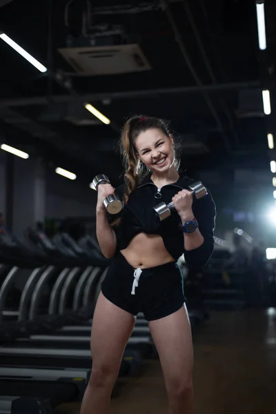 Fitness meisje poseren in de sportschool met halters in haar handen, haar lichaam pronken — Stockfoto