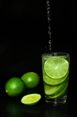 Yaz serinletici ve içecek konsepti. Su akışı içine soğuk içecek buz ve olgun dilim Yeşil taze limes bardak siyah arka plan üzerine dökülür. Ev yapımı limonata. Mojito kokteyl.