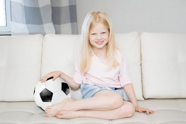 sport, people and entertainment concept - happy blonde girl with soccer ball sitting at home and watching football game on tv