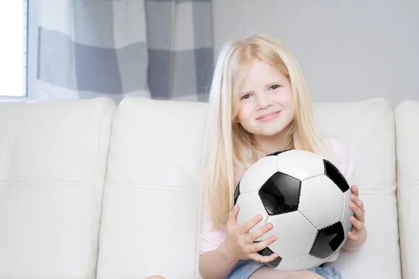 sport, people and entertainment concept - happy blonde girl with soccer ball sitting at home and watching football game on tv