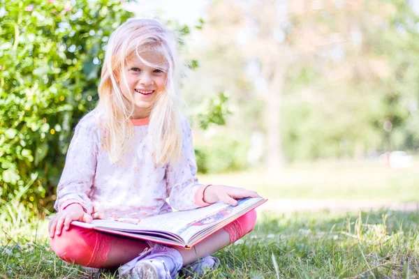 education, knowledge, and people concept - blonde prodigy little girl is reading big book in garden at summer day and smiling at camera