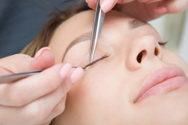 Beauty and fashion concept - Eyelash Extension Procedure. Woman face close up. Master of tweezers is working in salon