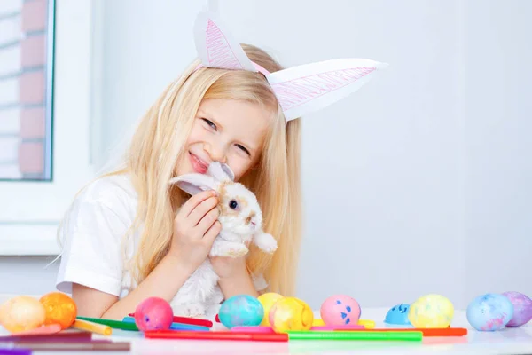 Blonde girl in rabbit ears on head and little bunny in her hands. Colorful eggs and markers on table. Prepearing for Easter and holidays concept.