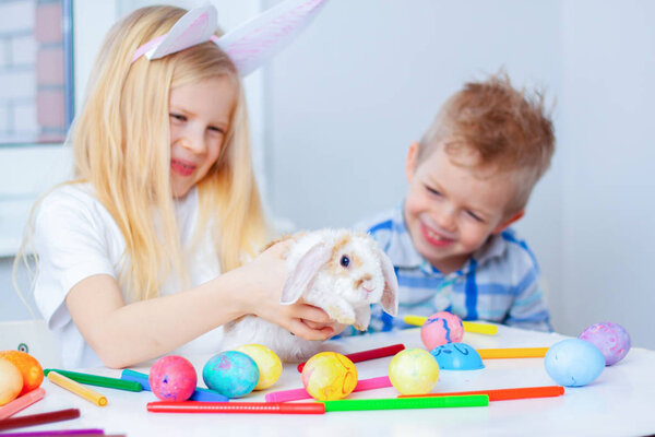 Блондинка в кроличьих ушах и мальчик с маленьким кроликом. Красочные яйца и маркеры на столе. Подготовка к Пасхе, семье и праздникам
