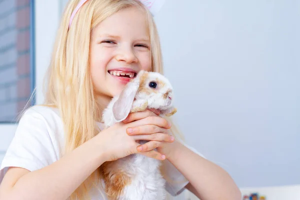 Blonde girl in rabbit ears on head and little bunny in her hands. Colorful eggs and markers on table. Prepearing for Easter and holidays concept