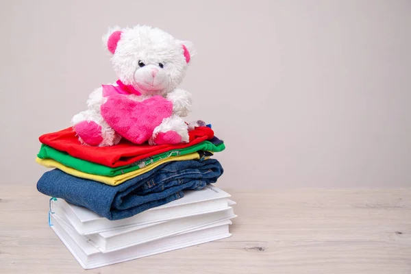 Donation concept. Donate goods with kids clothes, books, school supplies and toys. Teddy bear with big pink heart in hands. Copyspace for text.