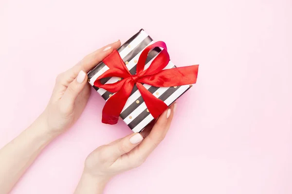 妇女手与礼物礼物箱子在白色和黑色条纹与红色丝带弓查出在粉红色的桌面视图。最低限度的平面组合销售, 生日, 母亲节或圣诞节。复制空间 — 图库照片