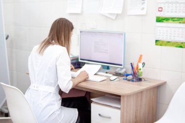 Klinik hastanede beyaz üniformalı beyaz üniformalı beyaz sarışın kadın doktorun arka yüzü bilgisayarda çalışıyor. Dolap ve arka planda diğer tıbbi ekipman. Sağlık kavramı